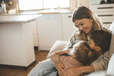 Smiling affectionate young woman embracing children sitting on sofa - JOSEF09488