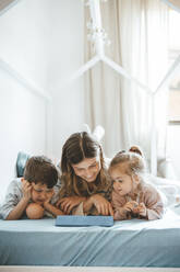 Lächelnde Frau mit Sohn und Tochter schaut auf einen Tablet-PC auf dem Bett liegend - JOSEF09497