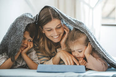 Smiling young woman and children under blanket looking at tablet PC lying on bed - JOSEF09498