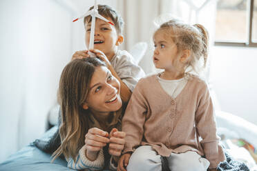 Playful boy holding wind turbine on mother's head sitting by sister at home - JOSEF09506