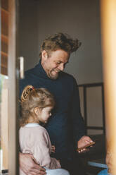 Happy man showing mobile phone to daughter seen through window - JOSEF09525