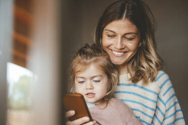 Smiling young woman showing mobile phone to daughter seen through window - JOSEF09530