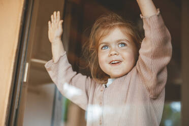 Glückliches süßes Mädchen zu Hause, das durch ein Glasfenster hinausschaut - JOSEF09533