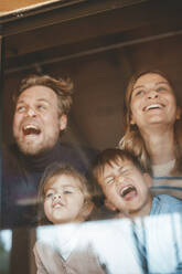 Playful parents and children pressing faces on window glass - JOSEF09536