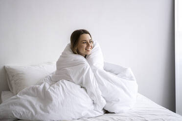 Happy woman wrapped in blanket sitting on bed at home - SSGF00916