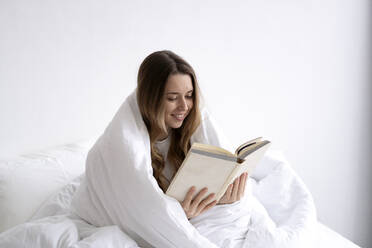 Smiling young woman wrapped in white blanket reading book on bed at home - SSGF00917