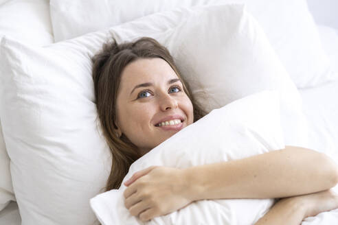 Happy woman embracing pillow lying on bed day dreaming at home - SSGF00924