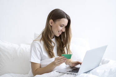 Happy woman holding credit card doing online shopping through laptop in bedroom at home - SSGF00931
