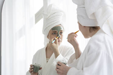 Frau mit Pinsel beim Auftragen einer Gesichtsmaske auf das Gesicht zu Hause - SSGF00937
