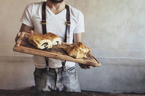 Baker holding cutting board with croissants and bread in front of wall - JUBF00396