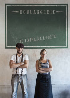 Confident bakery owners standing with arms crossed in front of wall with French text - JUBF00398