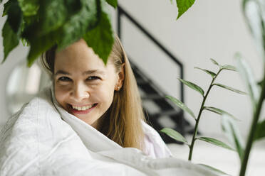 Lächelnde Frau in Decke eingewickelt bei Pflanzen zu Hause - SEAF00933