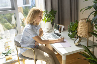Blond freelancer writing in dairy sitting on chair at desk in home office - SVKF00155