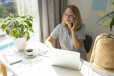 Lächelnde blonde Freiberuflerin, die am Schreibtisch im Heimbüro sitzt und mit ihrem Smartphone spricht - SVKF00160