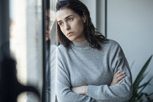 Depressive Frau steht mit verschränkten Armen und schaut durch das Fenster zu Hause - JSRF02001