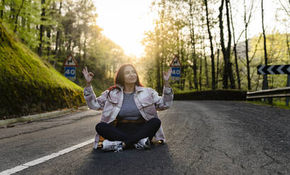 Smiling woman gesturing sitting cross-legged on road - JCCMF06272