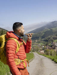 Mann mit Rucksack sendet an einem sonnigen Tag eine Sprachnachricht über sein Mobiltelefon - JCCMF06275