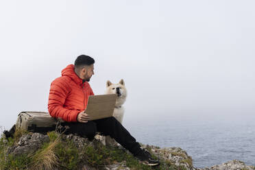 Mann sitzt mit Laptop und betrachtet seinen Hund auf einem Felsen - JCCMF06296
