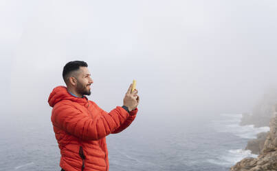 Lächelnder Mann, der mit seinem Smartphone fotografiert und vor dem Meer steht - JCCMF06297