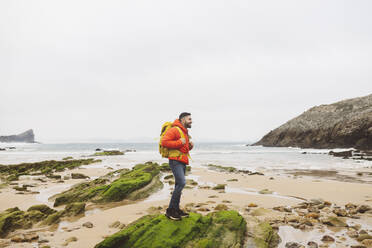 Mann mit Rucksack steht auf einem Felsen am Strand - JCCMF06304