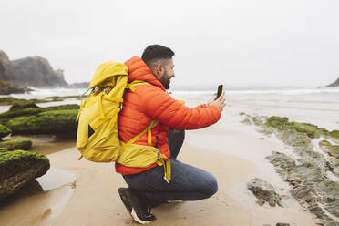 Man wearing backpack crouching photographing through smart phone at beach - JCCMF06311