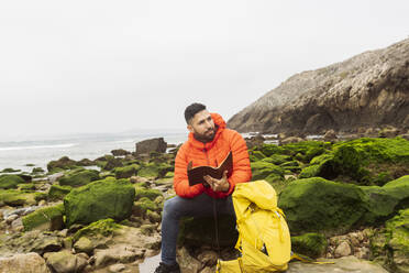 Man with diary and backpack sitting on rock at beach - JCCMF06312