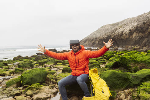 Glücklicher Mann mit Virtual-Reality-Simulator sitzt mit ausgestreckten Armen auf einem Felsen - JCCMF06313