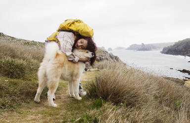 Smiling woman wearing backpack hugging pet dog - JCCMF06323