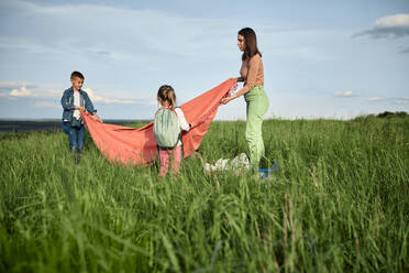 Mother and children laying out picnic blanket on grass in field - ZEDF04542