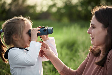 Daughter looking at mother through binoculars in field - ZEDF04553