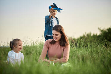 Smiling mother and daughter in front of boy playing with toy airplane on field - ZEDF04573