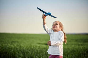 Playful girl flying airplane toy on field at sunset - ZEDF04578