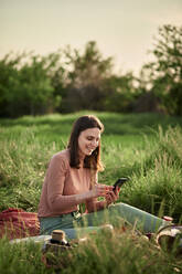 Lächelnde junge Frau, die beim Picknick auf einem Feld ihr Smartphone benutzt - ZEDF04584