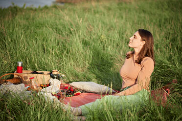 Junge Frau mit geschlossenen Augen auf einer Picknickdecke auf einem Feld sitzend - ZEDF04588