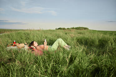 Young woman reading book lying on grass on field at sunset - ZEDF04590