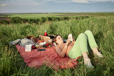 Smiling woman reading book at picnic in field - ZEDF04592