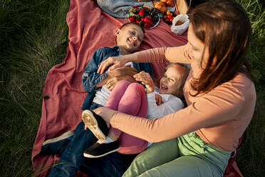 Mother tickling children lying on picnic blanket in field - ZEDF04601