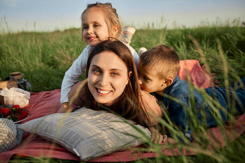 Smiling mother lying by children in picnic blanket at field - ZEDF04604