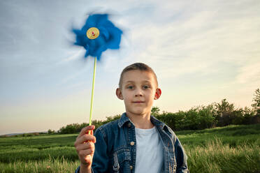 Niedlicher Junge hält blaues Windrad-Spielzeug auf einem Feld bei Sonnenuntergang - ZEDF04610