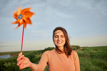 Lächelnde junge Frau, die ein Windrad mit geschlossenen Augen auf einem Feld bei Sonnenuntergang hält - ZEDF04615