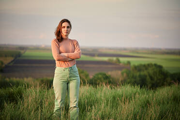 Selbstbewusste junge Frau mit verschränkten Armen auf einem Feld bei Sonnenuntergang stehend - ZEDF04626