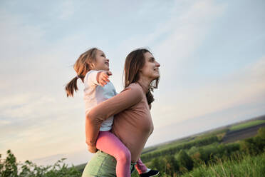 Glückliche Mutter nimmt ihre süße Tochter bei Sonnenuntergang im Feld huckepack - ZEDF04629
