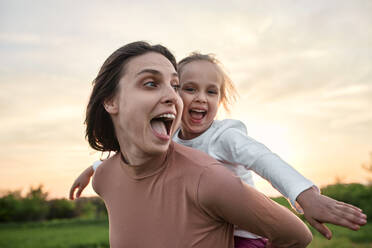 Cheerful mother carrying daughter piggyback in field on weekend - ZEDF04630