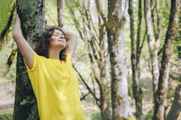 Carefree woman leaning on tree trunk in forest - OMIF00807