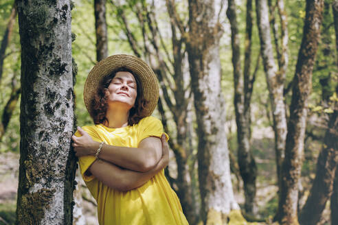 Frau mit Hut umarmt sich selbst an einem Baum an einem sonnigen Tag - OMIF00809