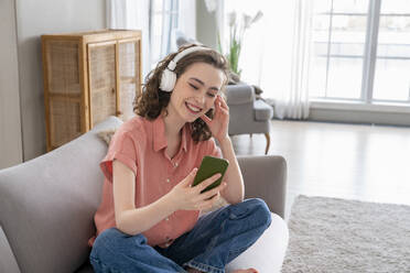 Happy woman wearing wireless headphones using smart phone on sofa at home - VPIF06049