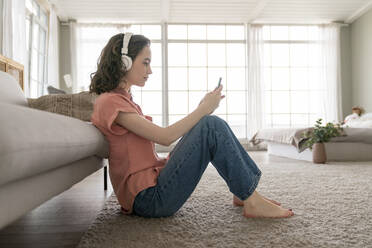 Young woman using smart phone sitting on carpet at home - VPIF06055