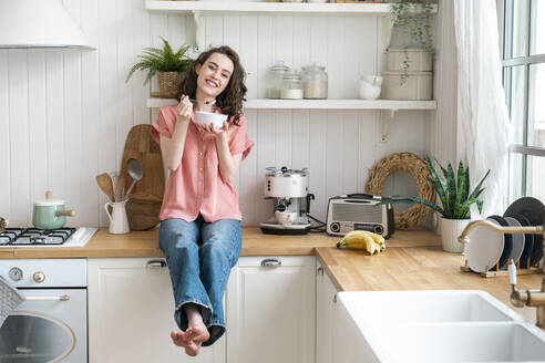 Happy woman eating breakfast sitting on kitchen counter at home - VPIF06058