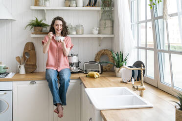 Happy young woman eating breakfast sitting on kitchen counter at home - VPIF06060