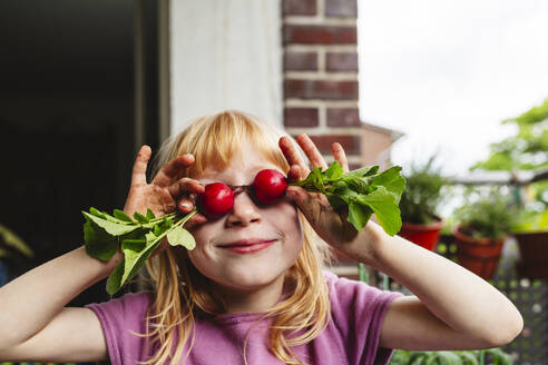Smiling girl covering eyes with fresh radish on balcony - IHF00833